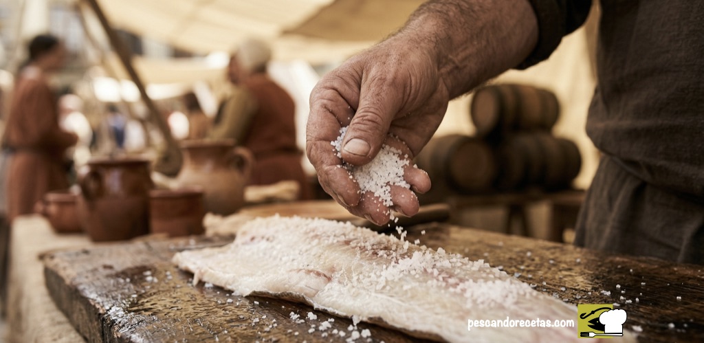 Primer plano de una mano curtida de un pescador vasco echando sal gorda sobre un lomo de bacalao abierto en un mercado medieval.